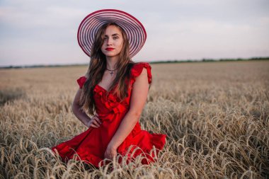 Genç, ince, parlak kırmızı elbiseli ve şapkalı seksi bir kız gün batımında buğday tarlasında duruyor. Yüksek kalite fotoğraf