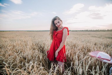 Genç, ince, parlak kırmızı elbiseli ve şapkalı seksi bir kız gün batımında buğday tarlasında duruyor. Yüksek kalite fotoğraf