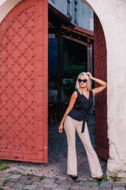 Sarışın, güneş gözlüklü, siyah bluzlu ve bej pantolonlu güzel bir kız eski bir yakanın arkasında poz veriyor. Yüksek kalite fotoğraf