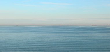 Deniz manzarası, mavi gökyüzü ve ortasında uzun yatay çizgi olan geniş mavi deniz açısı ve uzaktaki yol kenarında gemiler.