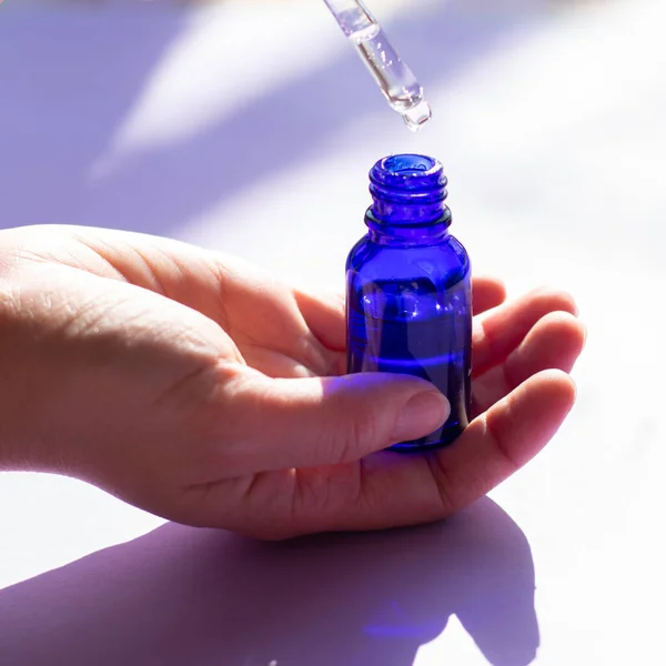 Yüz bakımıyla dolu mavi cam şişeli kare format. Tedavi için şeffaf sıvı serum. Spa prosedürü kozmetik ürünleri. Kadın elleri.