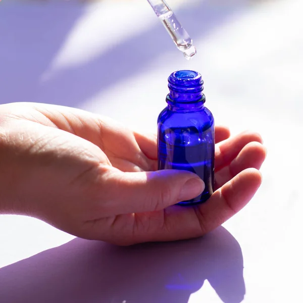 蓝色玻璃瓶的正方形 用满脸皮肤护理半透明液体血清进行治疗 温泉疗程化妆品 女性的手 图库图片