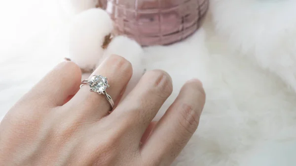 Close up of an elegant diamond ring on woman finger with pink perfume and white feather background. love and wedding concept. Soft and selective focus.