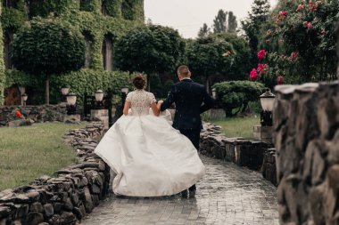 Gelin ve damat, yağmurda süzülen bembeyaz bir elbiseyle sarmaşıkla kaplanmış güzel bir binanın arka planına doğru koşuyorlar.