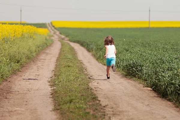 Kid having happy time running on road in the field of rapeseed — Stock Photo, Image