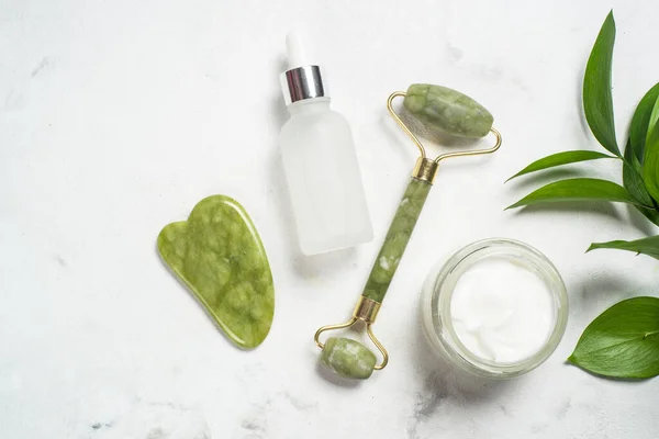 Jade rolo e água sha pedra massageador com creme facial e garrafa de soro. — Fotografia de Stock