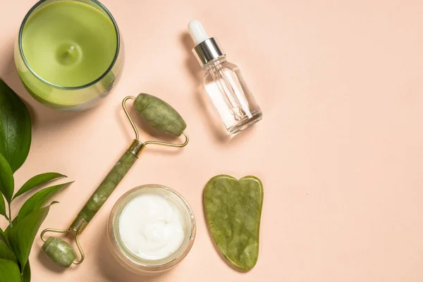 Kozmetik ürünleri - Yeşim silindirli ve kremalı gua sha masajcısı ve pastel arka planda serum şişeleri. — Stok fotoğraf