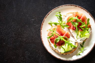 Krem peynirli ve jambonlu sandviç ya da taze yeşillikli beyaz tabak. Koyu masadaki üst görünüm.