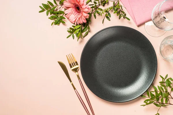 Siyah tabak, bardaklar, çatal bıçak takımı ve pembe arka planda çiçekler. Kopyalama alanı olan üst görünüm.