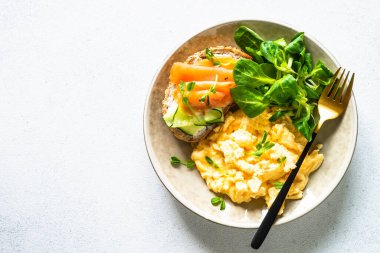 Sağlıklı kahvaltı ya da öğle yemeği. Çırpılmış yumurta, krem peynirli sandviç, somon balığı ve beyaz taş masada salata yaprakları. Kopyalama alanı olan üst görünüm.