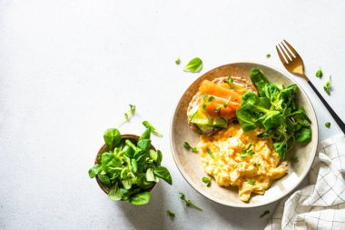 Sağlıklı kahvaltı ya da öğle yemeği. Çırpılmış yumurta, krem peynirli sandviç, somon balığı ve beyaz taş masada salata yaprakları. Kopyalama alanı olan üst görünüm.