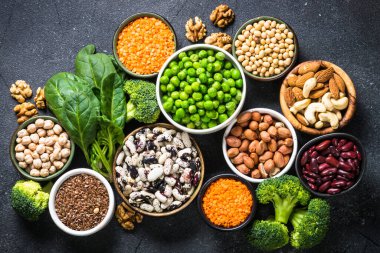 Vegan protein kaynağı, sağlıklı beslenme gıdası. Baklagiller, fasulyeler, fındıklar, sebzeler ve tohumlar. Siyah taş masadaki üst görünüm.