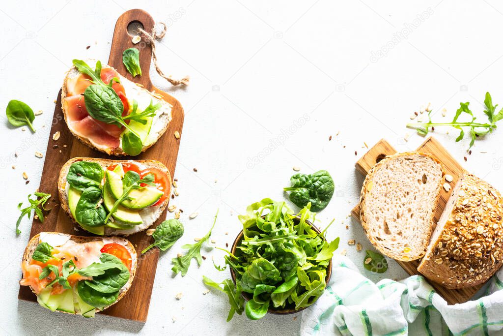 Open sandwiches set with cream cheese, prosciutto, salmon, avocado and fresh greens and vegetables. Top view at white table with copy space.