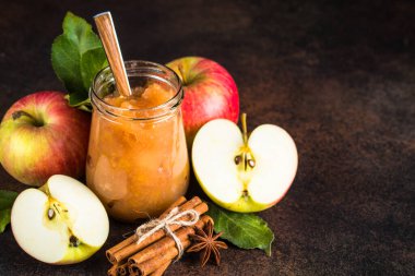 Cam kavanozdaki baharatlı elma reçeli ve koyu taş zemin üzerinde taze elmalar..