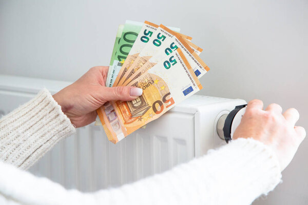 Экономия отопления, дорогостоящая оплата отопления. Банкноты с деньгами от радиатора и евро в руках женщины.
