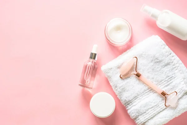 Natural cosmetics on pink. Skin care product, cream, soap serum, jade roller and white towel. Flat lay image with copy space.