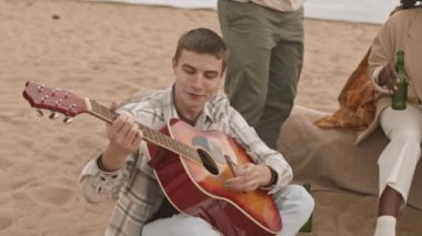 Yüksek açılı, orta boylu, akustik gitar çalan beyaz bir adam. Bulutlu bir günde kumlu bir sahilde, kesilmiş arkadaşlarıyla oturuyor.