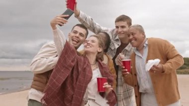 Orta uzunlukta beyaz bir kadın, plajda elinde kırmızı plastik bardaklarla mutlu çok ırklı arkadaşlarıyla selfie çekmek için akıllı telefon kullanıyor.