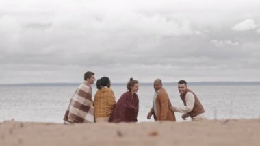 Sahilde dikilen, sarılan, battaniyelerle ısınan, soğuk bulutlu bir günde deniz manzarasının tadını çıkaran, arkasını dönüp kaçan ve gülümseyen beş genç insan.