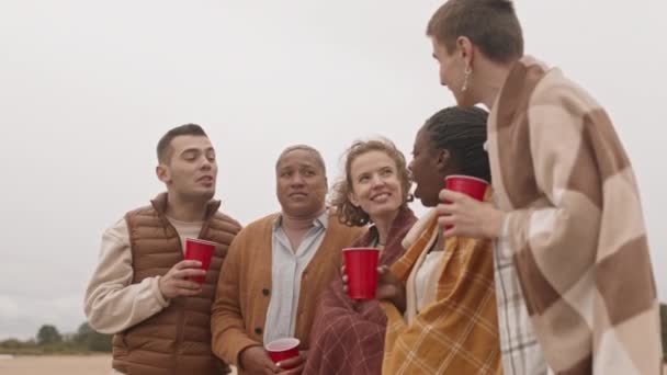 中长五个多民族的男女朋友站在海滩上 举着红色的塑料杯 在阴冷的天气里用毛毯热身 — 图库视频影像