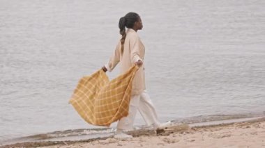 Genç Afrikalı bir kadının kareli atkısını tutarken, bulutlu bir günde kumlu deniz kıyısında yürürken.