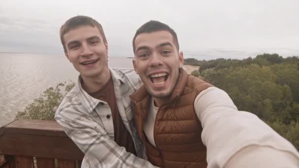 两名年轻白人男子手持Pov视频呼叫 并在镜头前微笑 展望海滨和灰蒙蒙的天空 — 图库视频影像
