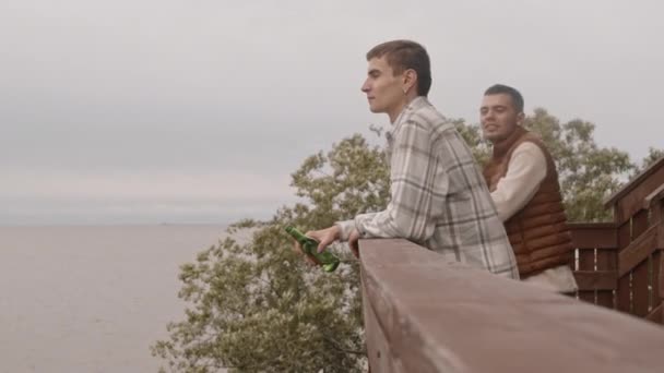 被困住了的年轻的白人男子站在夏宫的阳台上 他的不同的朋友和他在一起 手里拿着啤酒瓶 谈着话 面带微笑 — 图库视频影像