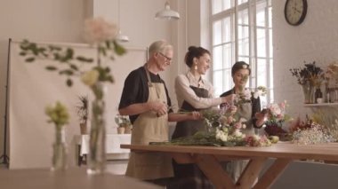 Çiçekçilerden oluşan neşeli bir ekip, çiçek buketleri düzenliyor ve küçük rahat bir çiçek stüdyosunda sohbet ediyorlar. Ön plandaki cam vazodaki güzel pembe karanfilin odak noktasını değiştiriyorlar.