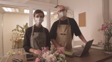 Kafkasyalı yetişkin çiftin küçük çiçek dükkanı sahibi maskeli, akıllı telefon ile ödeme yapan müşterilere güzel buket satan orta büyüklükteki portresi.