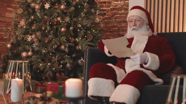 サンタクロースの中長い赤と白の衣装や眼鏡を着て 自宅でクリスマスツリーでアームチェアに座って 手紙を読んで笑顔 — ストック動画