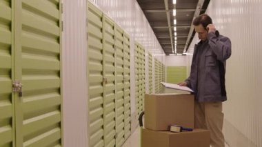 Orta uzunlukta beyaz erkek teslimat işçisi iş kıyafeti giyiyor, ambarın koridorunda karton kutuların yanında duruyor, panodaki kağıda bakıyor.