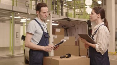 Orta uzunlukta beyaz kadın yönetici ve erkek kurye işçi tulumları giyer, kutularla dolu teslimat kamyonunun önünde durur, ayrılmadan önce konuşurlar.