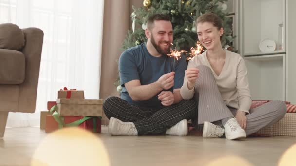 リビングルームで装飾されたクリスマスツリーの下に床に座っている幸せな若い白人の配偶者の広い ベンガルの光を保持します 笑顔とキス — ストック動画