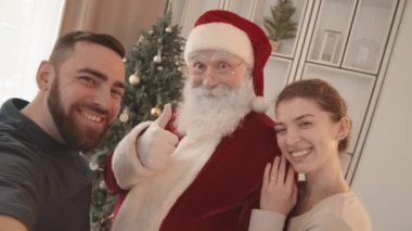Mutlu Noel Baba 'yla video çeken, el sallayan, gülümseyen ve oturma odasında ayakta duran beyaz kadın ve adamın el bilgisayarı.