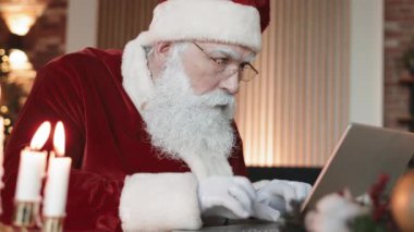 Beyaz sakallı Noel Baba 'nın gözlük taktığı, masasında oturduğu, dizüstü bilgisayarda yazdığı, sonra şaşırdığı ve kameraya baktığı orta boy yakın çekim.