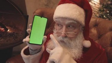 Noel Baba 'nın şapkası ve gözlüğü ile şöminenin yanındaki koltukta otururken, akıllı telefonu yeşil ekran ile tutarken, kameraya bakıp gülümserken.