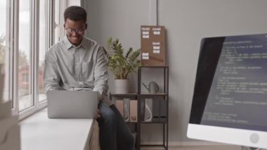 Genç Afro-Amerikalı erkek programcının dizüstü bilgisayarlı program kodlarıyla pencerenin kenarında otururken orta ölçekli bir fotoğrafı modern ofiste monitör ekranında görünüyor.