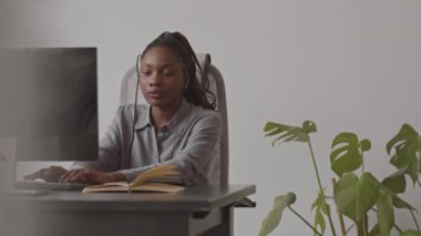 Afrika kökenli ciddi bir kadın programcının belden yukarı çekimi işyerinde oturmuş, bilgisayar monitörüne bakarken klavyede yazı yazıyor, deftere not alıyor.