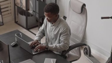 Çağdaş ofisteki iş yerinde otururken dizüstü bilgisayarında daktilo kodları yazan konsantre genç erkek Afro-Amerikalı programcının yüksek açılı görüntüsü.