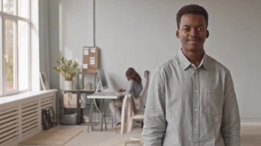 Kameraya göz kırpma için poz veren neşeli Afro-Amerikan erkek programcının orta boy bıçaklama portresi, modern bilişim ofisinde dururken, arka planda bilgisayar üzerinde çalışan bayan meslektaşı