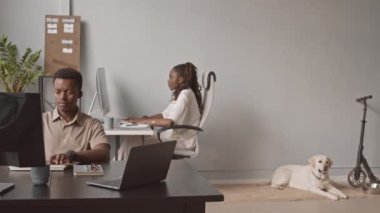 Genç Afro-Amerikalı kadın ve erkeğin masalarda oturup bilgisayarlarda yazışırken modern hayvan dostu ofiste çalışırken ve sevimli Labrador 'un e-scooter' ın yanında dinlenirken çekilmiş fotoğrafı.