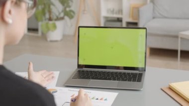 Genç iş kadınının yavaş çekimde dizüstü bilgisayarında pazarlama istatistiklerini tartışırken evden modern minimalist iç mekânla çalışırken videolu görüntüsü ile omzunun üstünden çekilmiş.