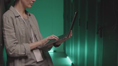 Orta uzunlukta dizüstü bilgisayarı olan, sunucu rafları arasında duran ve geriye yaslanan bir kadın bilişim uzmanı.