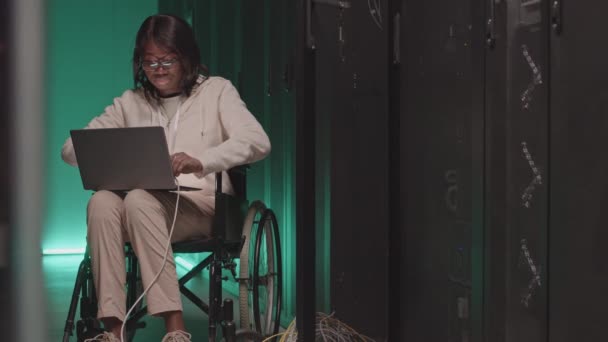 車椅子のアフリカ系アメリカ人女性の中長期 緑の照明データセンターでラップにポータブルコンピュータを保持し サーバキャビネットのワイヤ接続を扱う — ストック動画