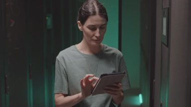 Tablet bilgisayar kullanan, yeşil ışıklı veri merkezinde duran ve sunucu kabinesinde yazı yazan beyaz tenli genç bir siber güvenlik uzmanı.