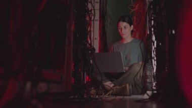Koyu renk saçlı beyaz kadın bilişim uzmanı taşınabilir bilgisayar kullanıyor, kırmızı ışıklı sunucu odasında birden fazla kablo arasında oturuyor.