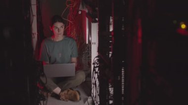 Koyu saçlı beyaz bir kadının taşınabilir bilgisayar kullanırken, kırmızı ışıklı sunucu odasında birden fazla kablo arasında otururken geniş açılı görüntüsü.