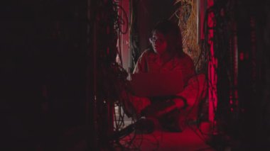 Genç Afrikalı Amerikalı bir kadının taşınabilir bilgisayar kullanırken, kırmızı ışıklı sunucu odasında birden fazla kablo arasında otururken geniş açılı görüntüsü.