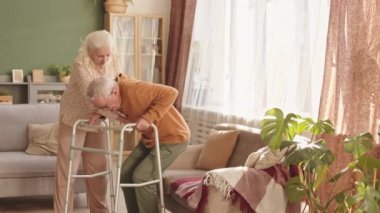 Orta boylu beyaz bir adam oturma odasında Walker 'ı kullanıyor, kanepeye oturuyor, destekleyici beyaz saçlı karısı ona yardım ediyor.