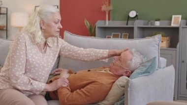 Orta boylu, beyaz saçlı bir kadının, burnunda oksijen tüpü olan yaşlı bir kocayla koltukta oturması, adamın karısını okşaması.